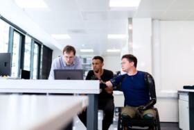 Three men looking at a laptop, one is a wheelchair user