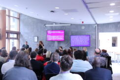 A room full of people watching a presentation. A man stands at a lectern, with an ISL interpreter beside him. There are screens behind them both, and other presenters sit facing the audience